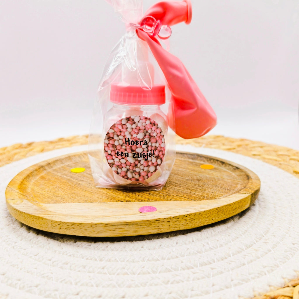 Geboorte traktatie meisje: roze babyflesje gevuld met roze en witte spekjes in cellofaan met lint, speentje en roze sticker.
